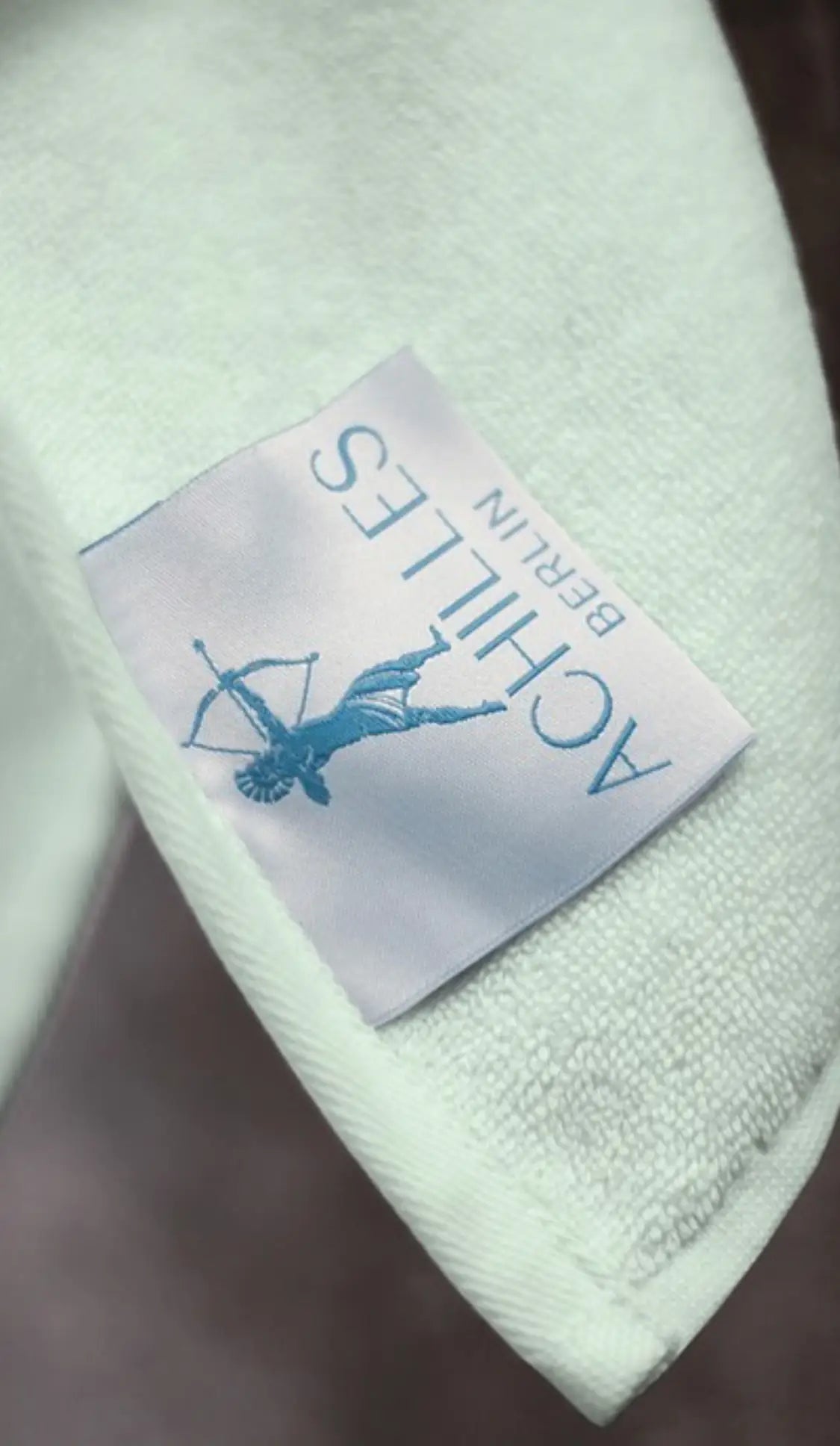 "Light blue cotton label with 'ACHILLES BERLIN' text and an illustration of a bowman, sewn onto a white fabric."