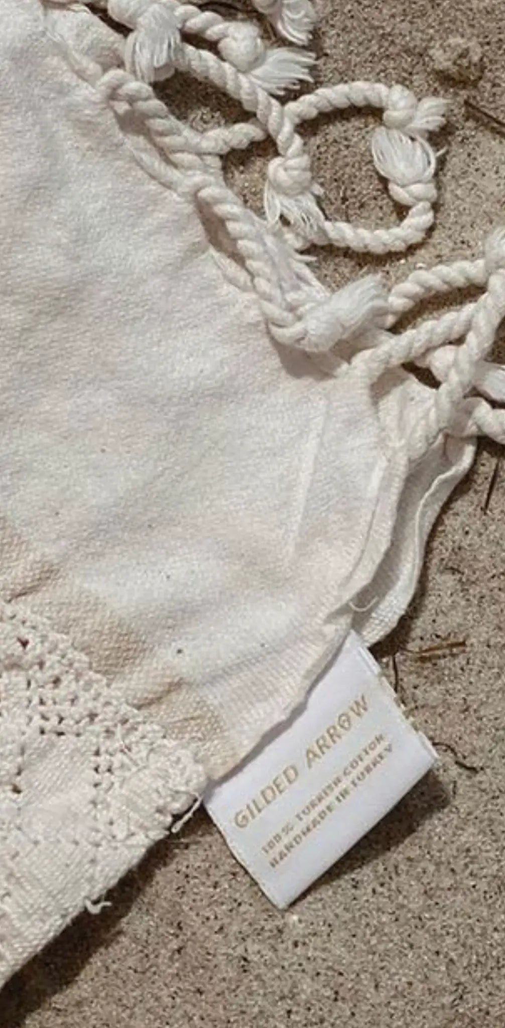 White cotton label with "GILDED ARROW" printed in black, attached to a textured fabric with braided tassels.