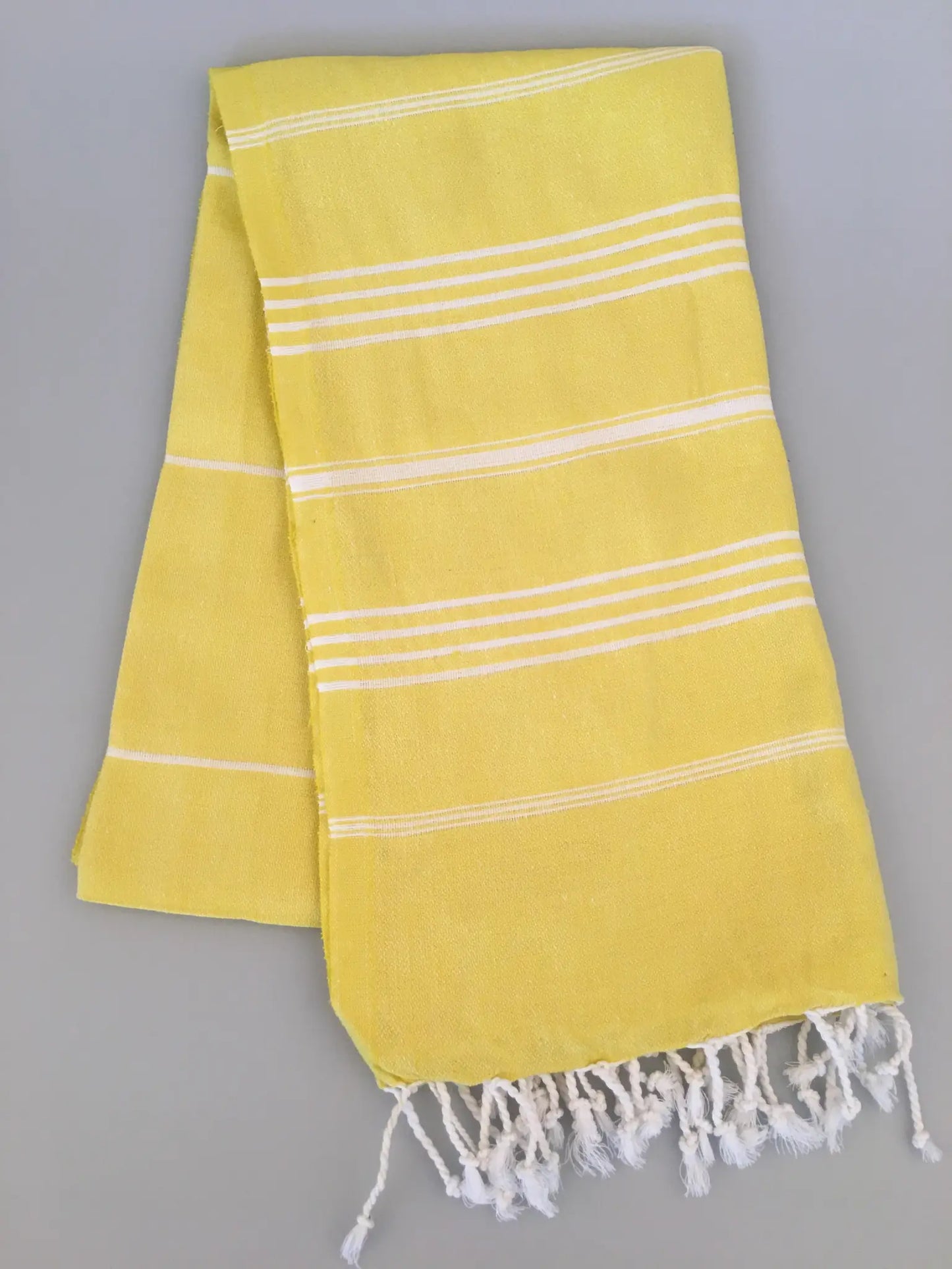 Yellow Turkish towel with white stripes and fringed edges, laid flat against a gray background.