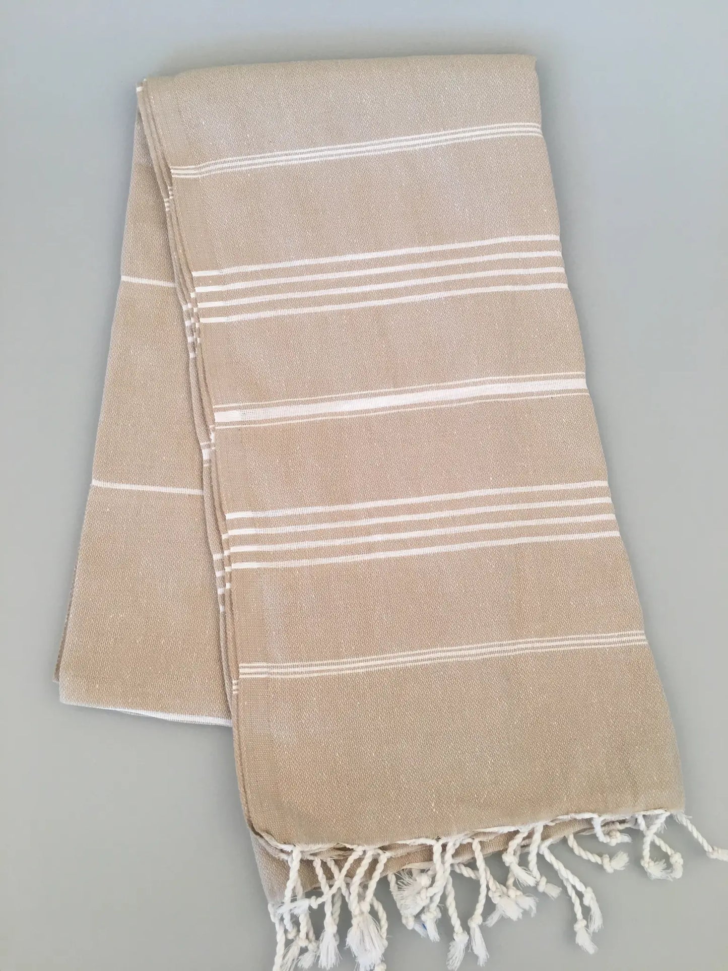 Beige Turkish towel with white stripes and fringed edges, folded flat on a light gray background.
