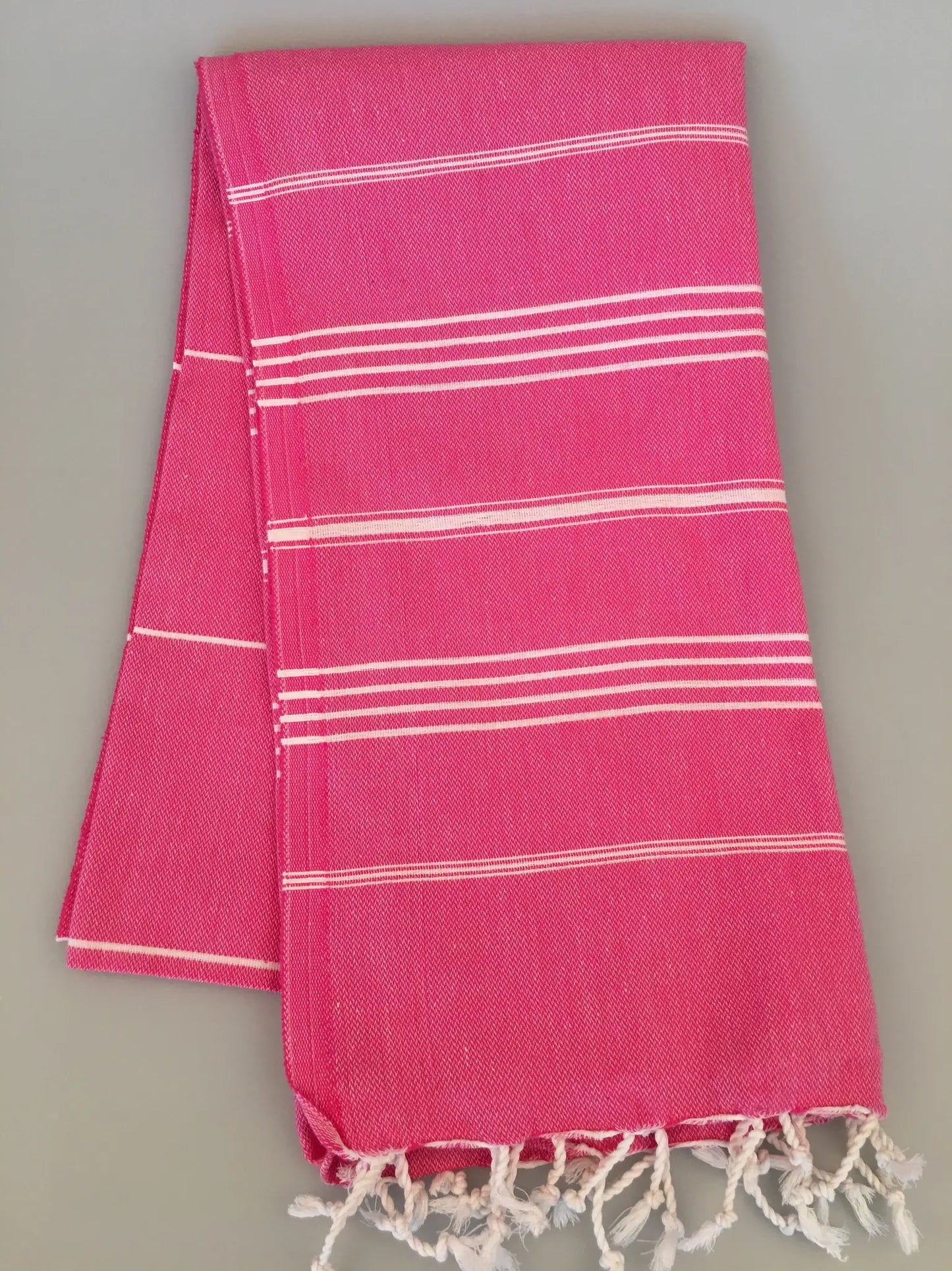 "Pink Turkish towel with white stripes and fringed edges, folded neatly on a gray background."