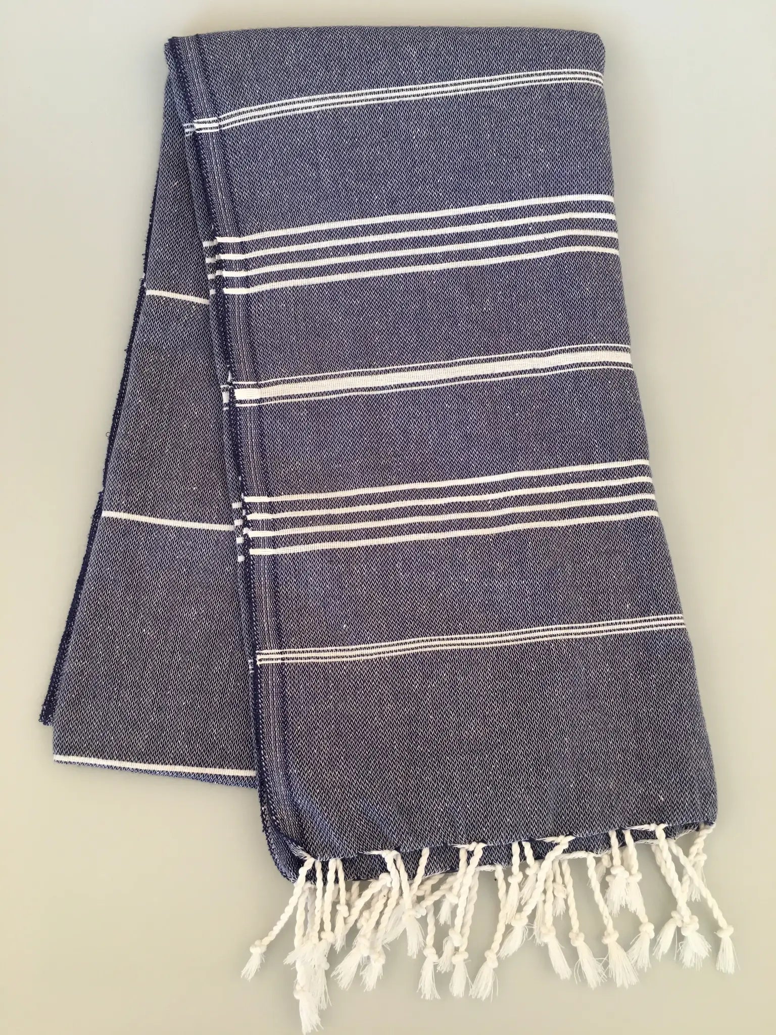 "Dark blue peshtemal towel with white stripes and fringe edges, neatly folded on a light background."
