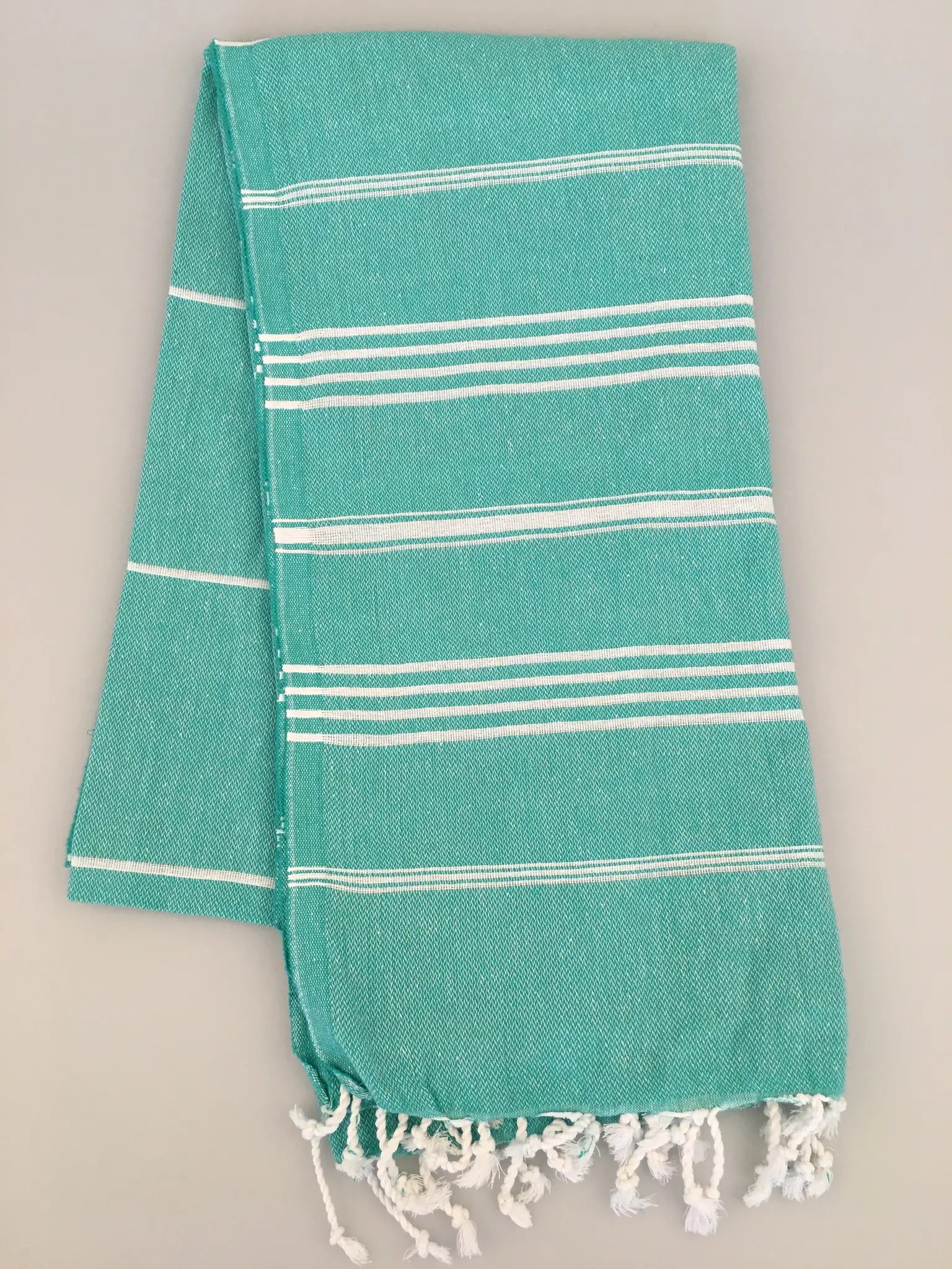 Turquoise Turkish towel with white stripes and fringed ends, laid flat on a light gray background.