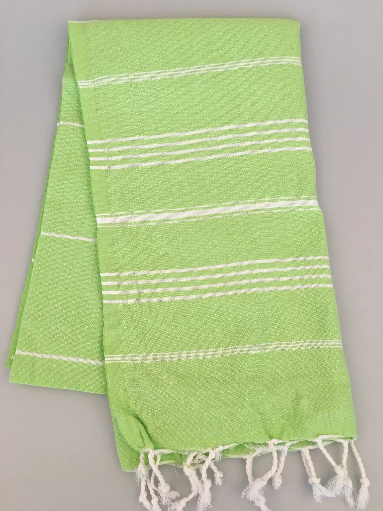 Light green Turkish towel with white stripes and fringe edges, folded neatly on a gray background.