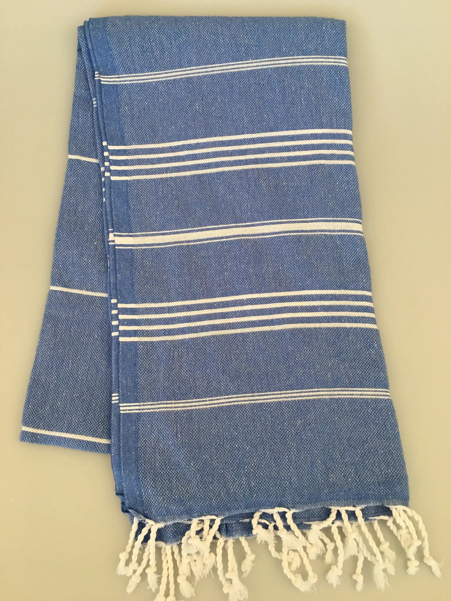 "Blue Turkish towel with white stripes and fringed edges, folded neatly on a light background."