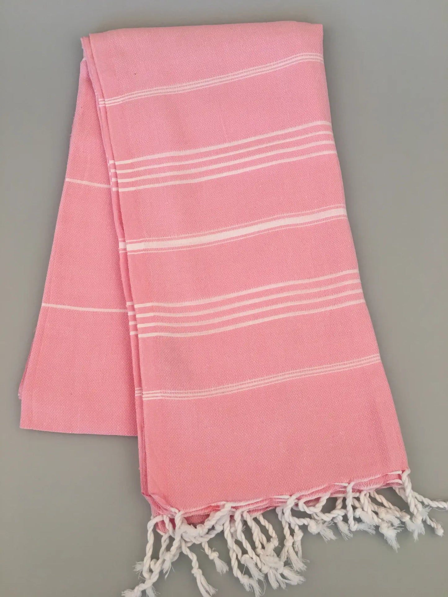 "Light pink Turkish towel with white stripes and fringed edges, laid flat on a gray surface."