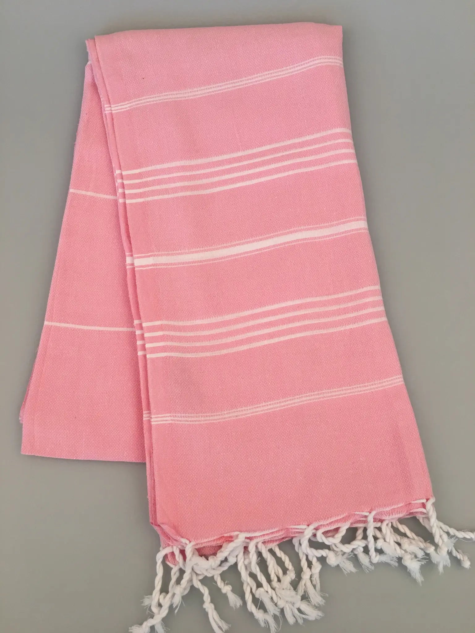 "Light pink Turkish towel with white stripes and fringed edges, laid flat on a gray surface."