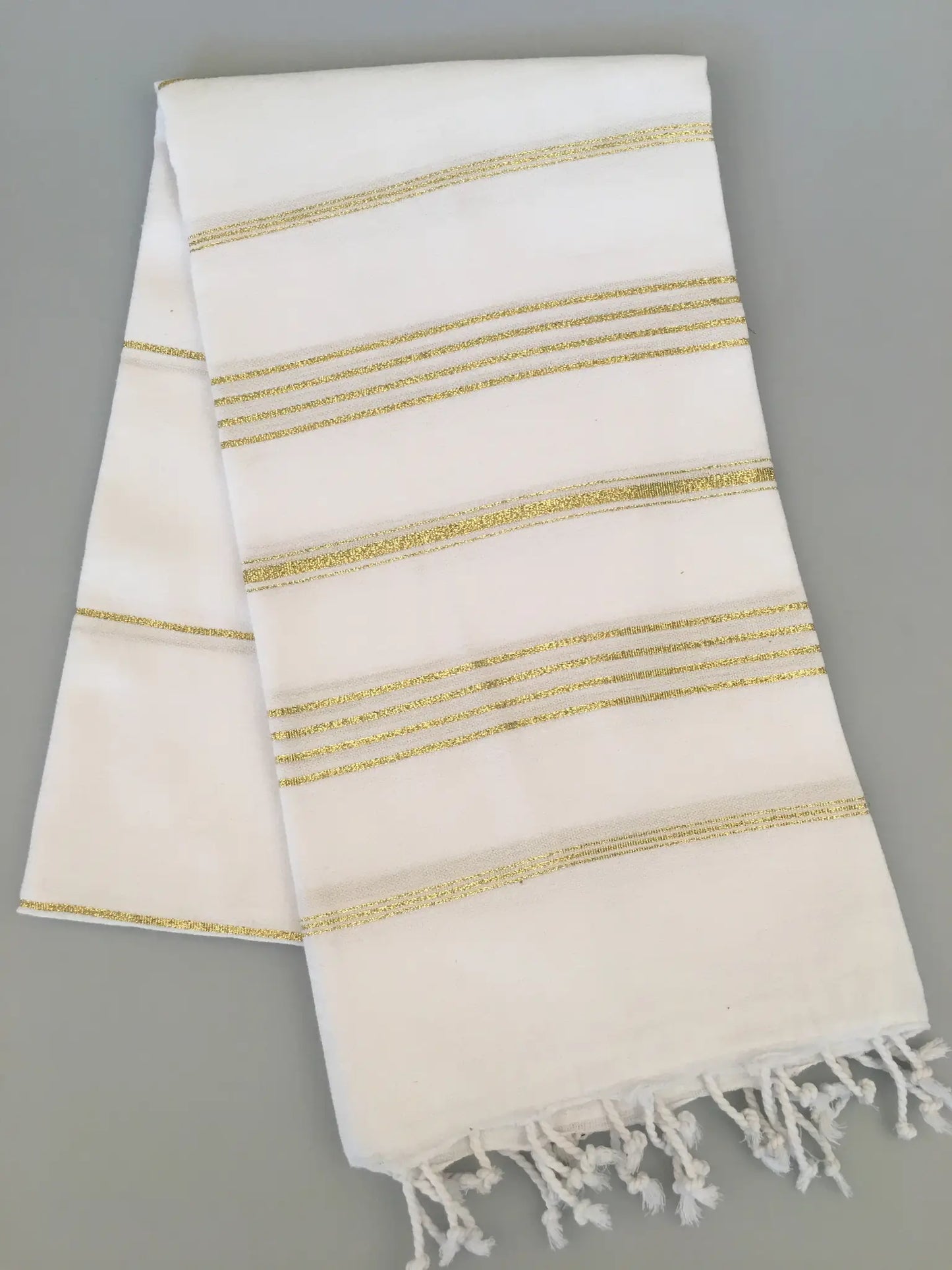 "White Turkish towel with gold striped pattern and fringed edges, folded on a gray background."