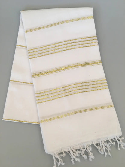 "White Turkish towel with gold striped pattern and fringed edges, folded on a gray background."
