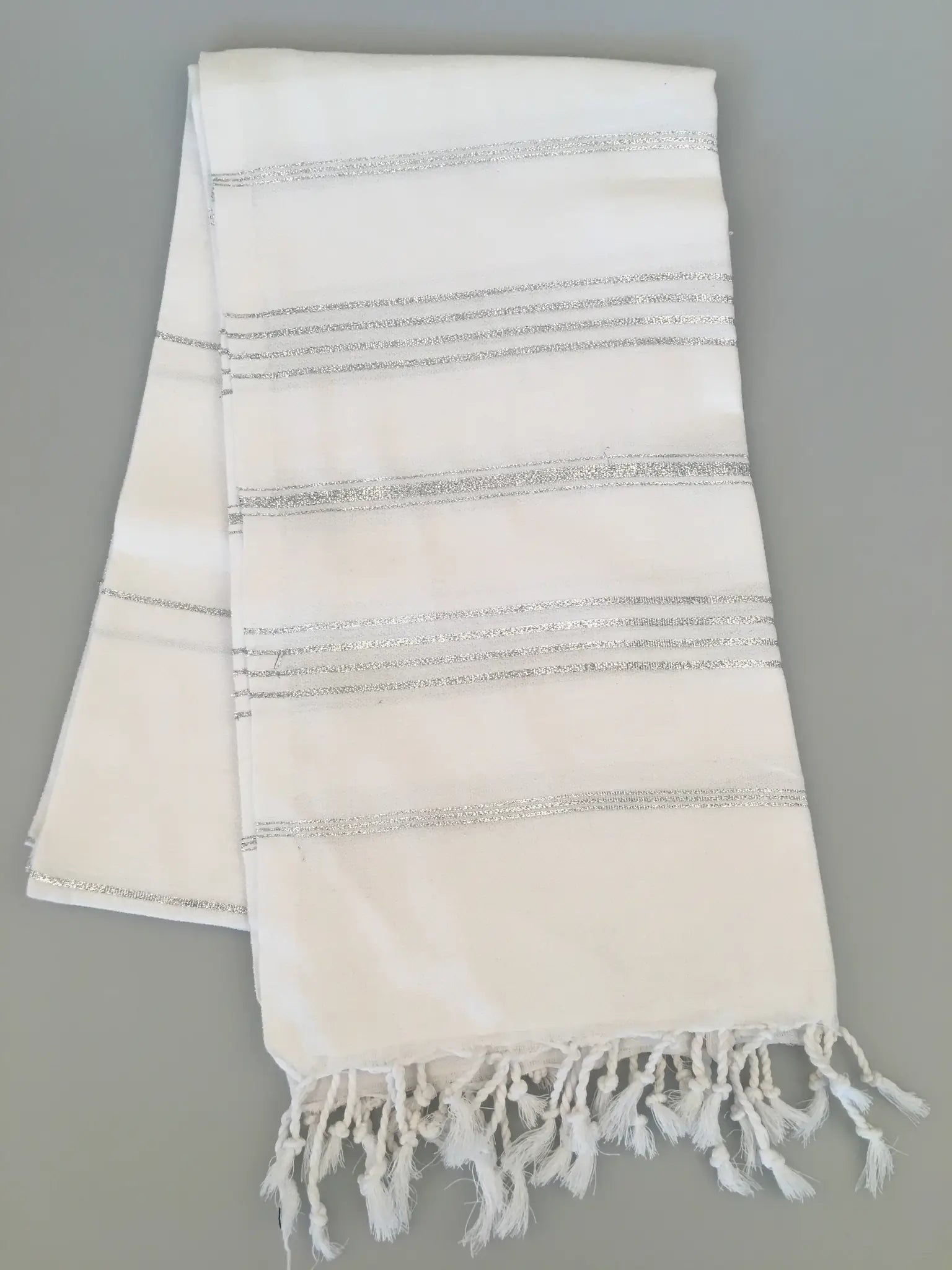 "White Turkish towel with silver striped patterns and fringed edges, laid flat on a light gray background."