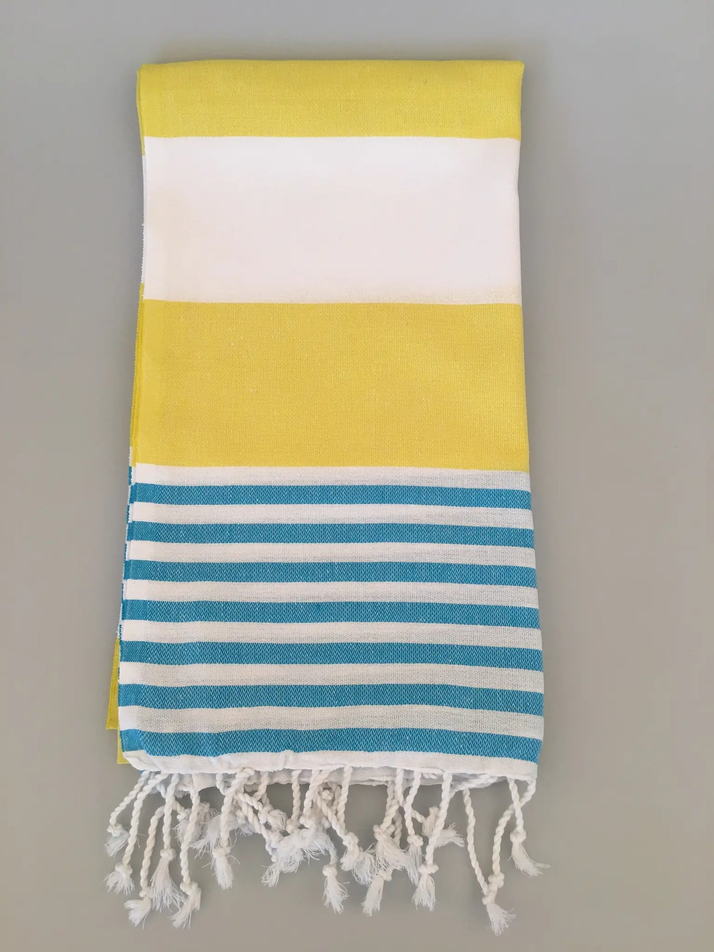 "Yellow and white striped Turkish towel with blue stripes at the bottom and fringed edges, folded flat."