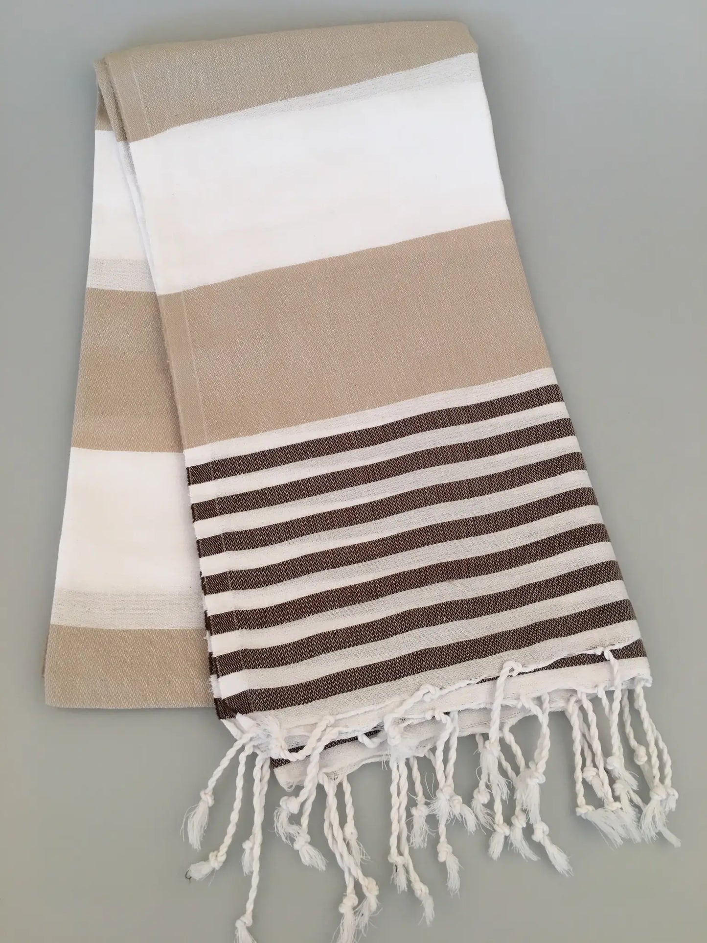 "Beige and white striped Turkish towel with fringe edges, folded on a light gray surface."