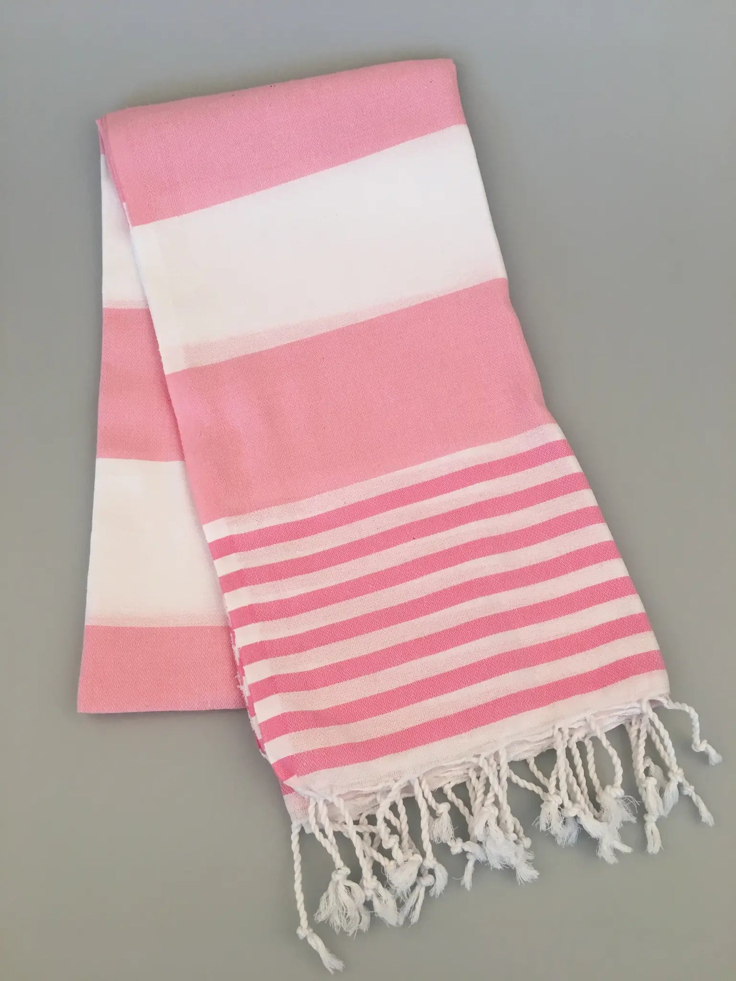 "Pink and white striped Turkish towel with fringed edges, folded on a light gray background."