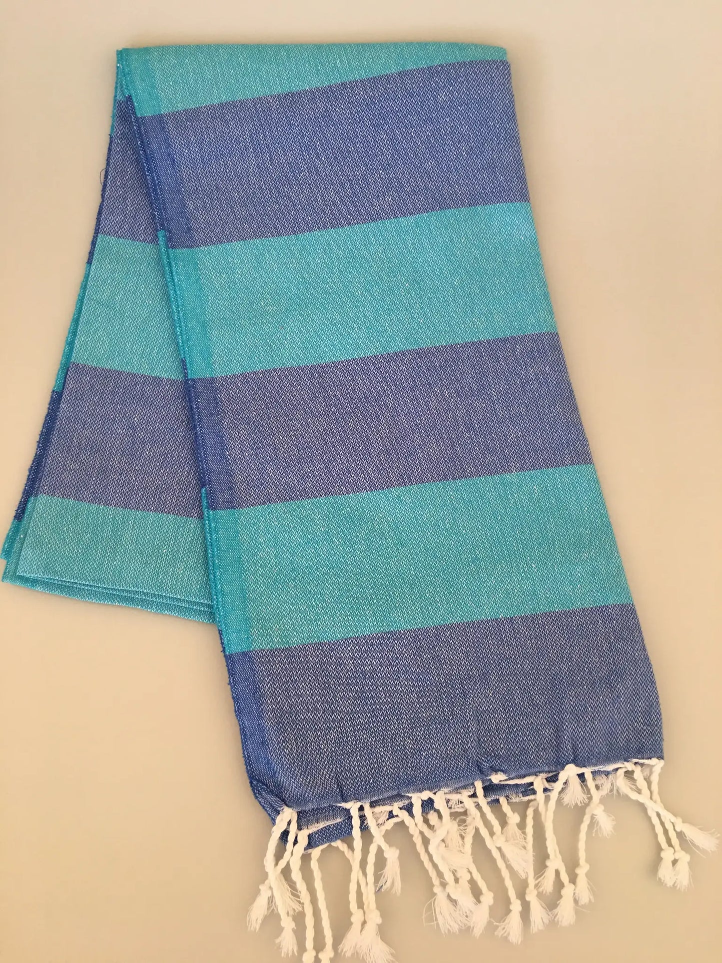 Turquoise and blue striped Turkish towel with fringed edges, laid flat on a light background.