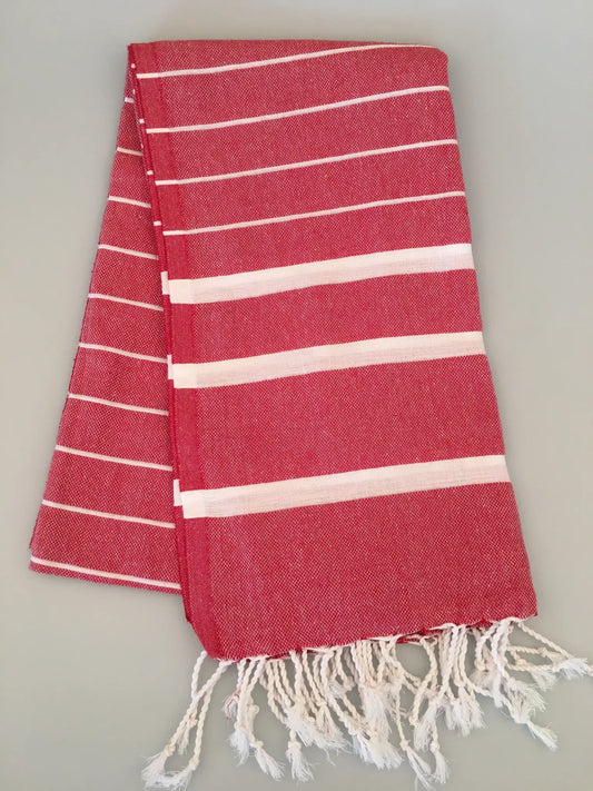 Red Turkish towel peshtemal with white stripes and fringed edges, folded neatly on a light gray background.
