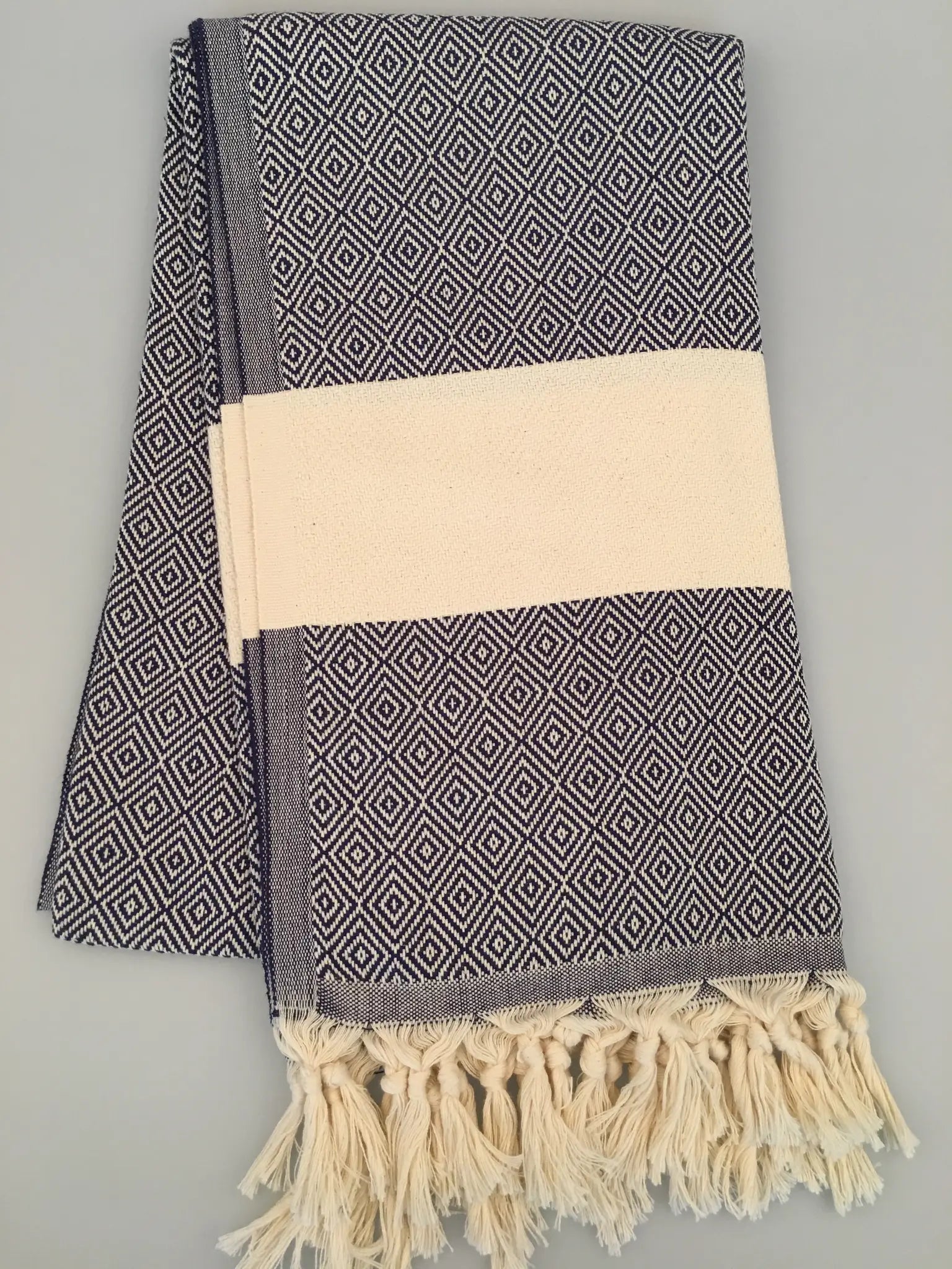 "Navy blue and white Turkish towel with diamond pattern, fringed edges, neatly folded with a cream band around it."