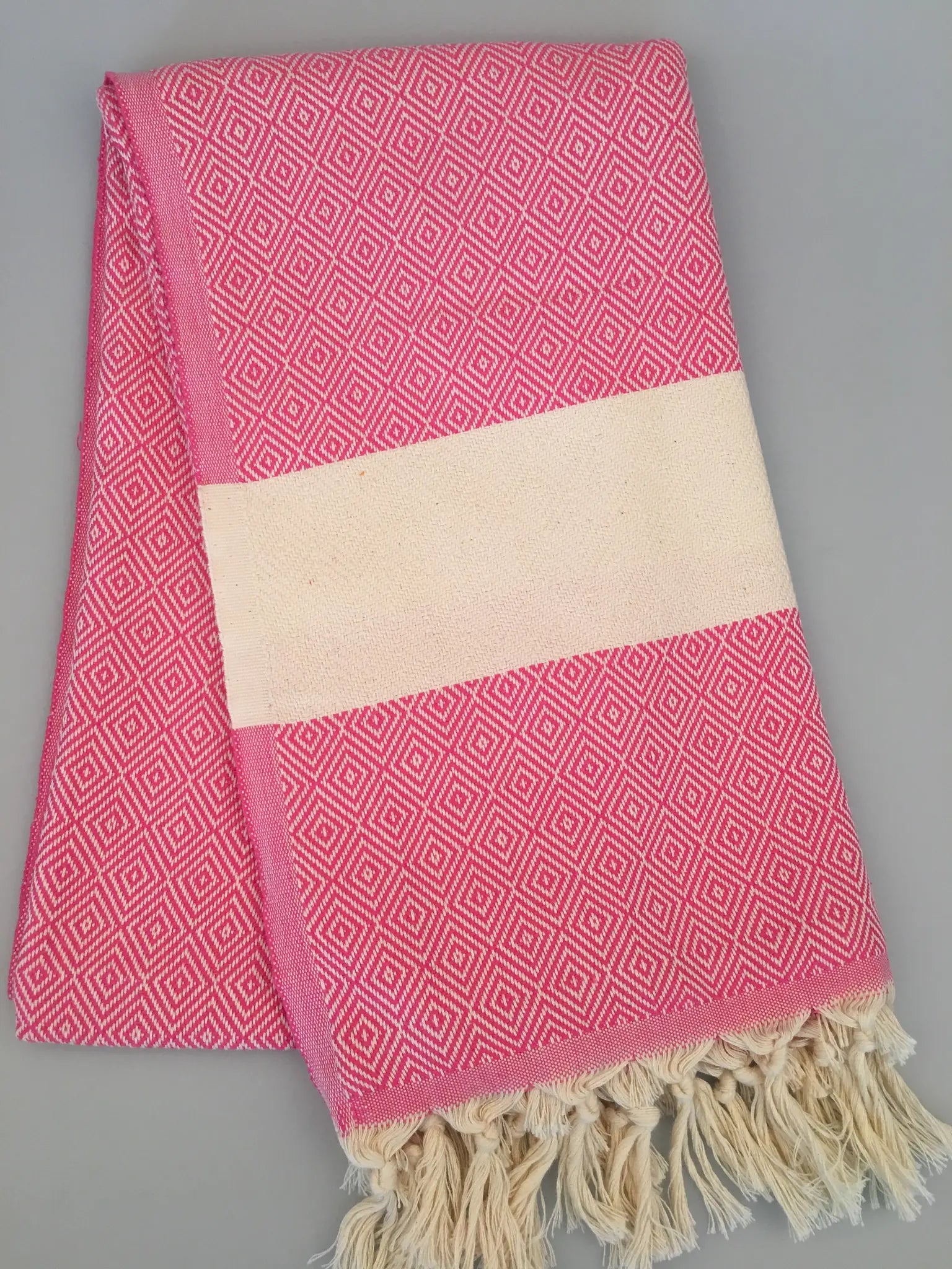 Pink Turkish peshtemal towel with diamond pattern, cream stripe in the center, and fringed edges, laid flat.