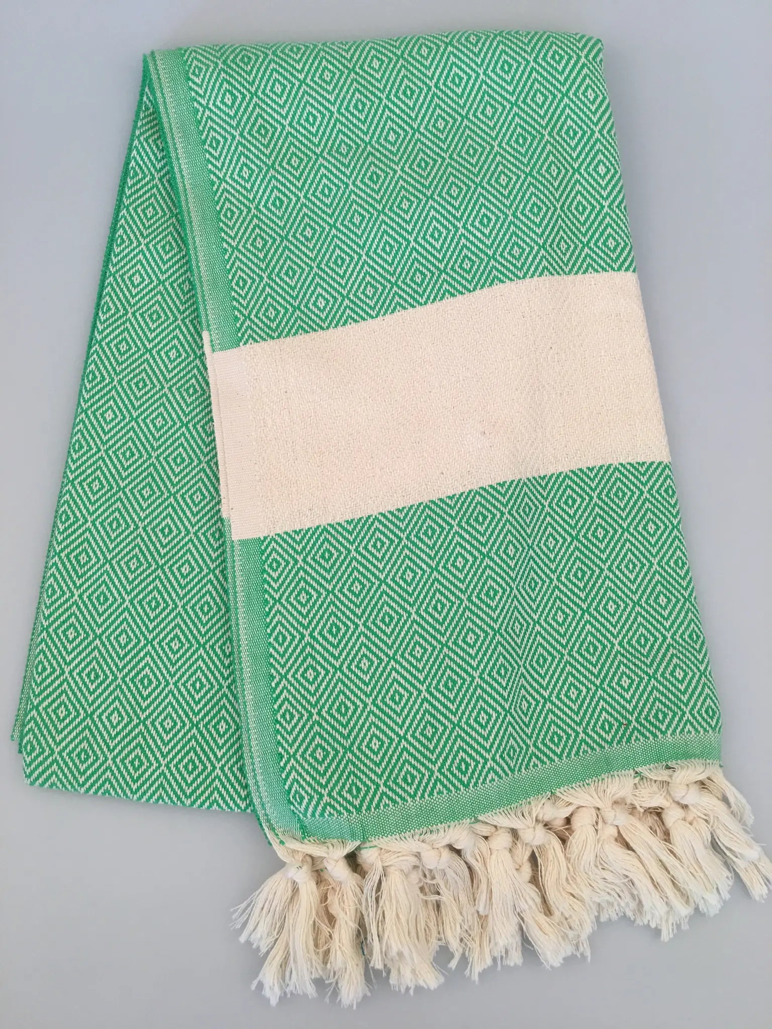 Green Turkish towel with diamond pattern, cream stripe, and fringed edges, neatly folded on a light gray background.