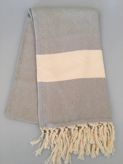"Gray Turkish towel with diamond pattern, cream-colored center band, and fringed edges, laid flat on a light surface."