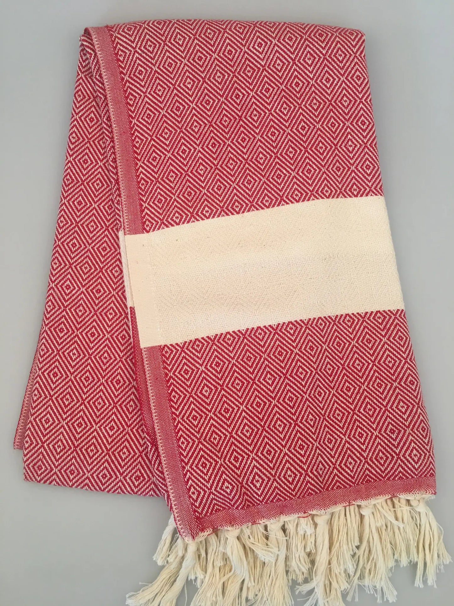 "Red Turkish towel with diamond pattern, cream band in the center, and fringed edges, laid flat on a gray background."