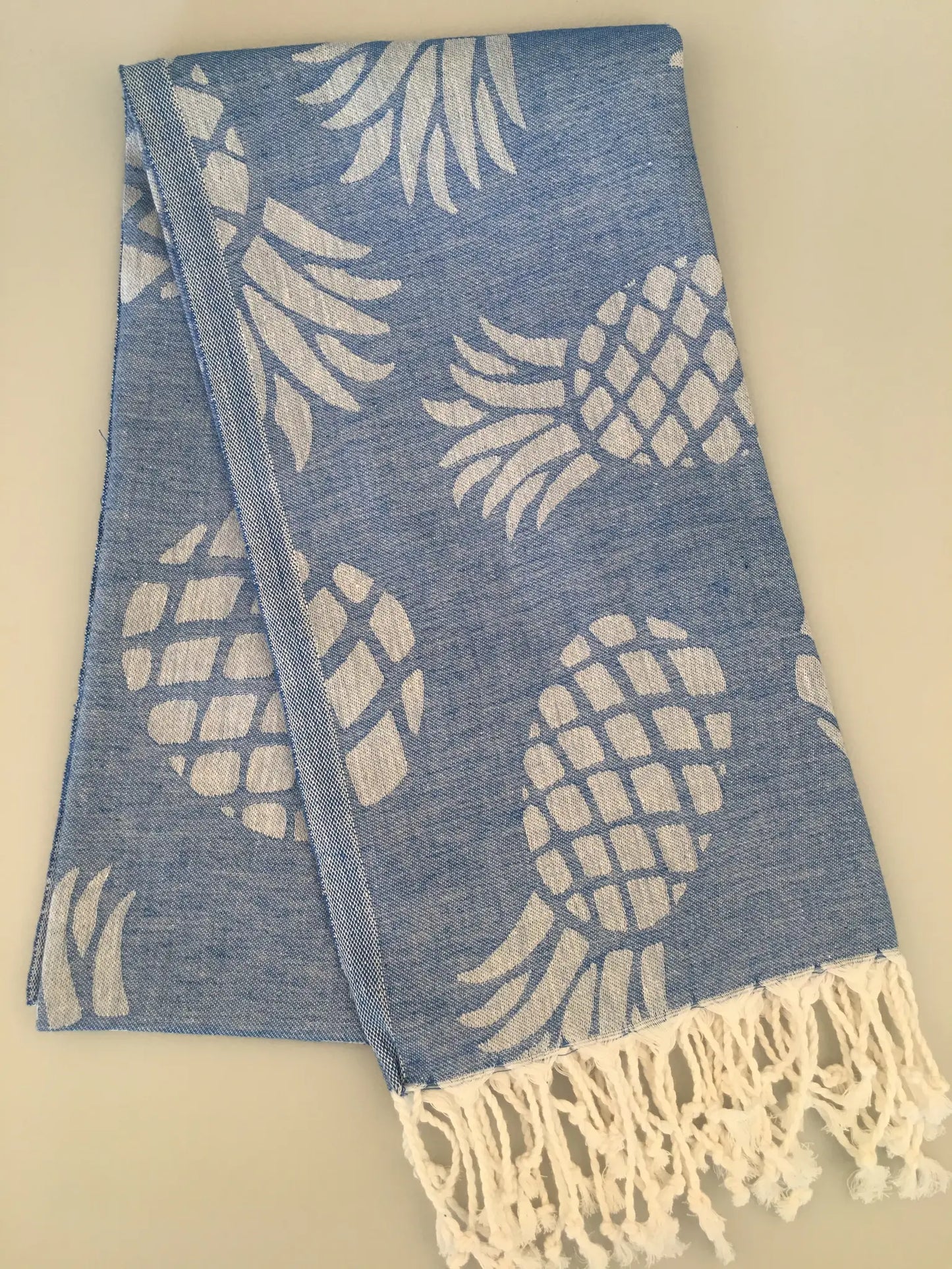 Blue Turkish towel with white pineapple jacquard pattern and fringed edges, folded on a light background.