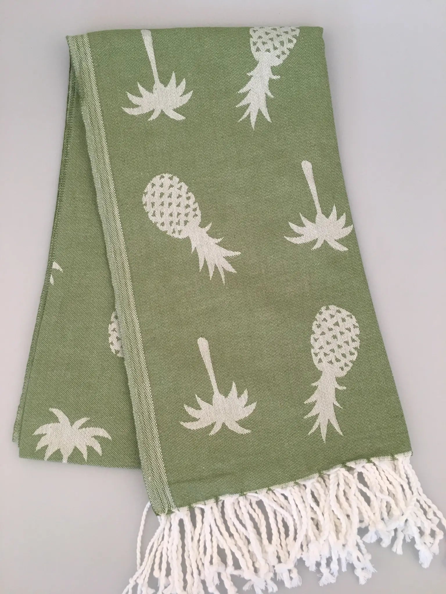 Green Turkish towel featuring white pineapple jacquard pattern and fringed edges, laid flat on a light background.