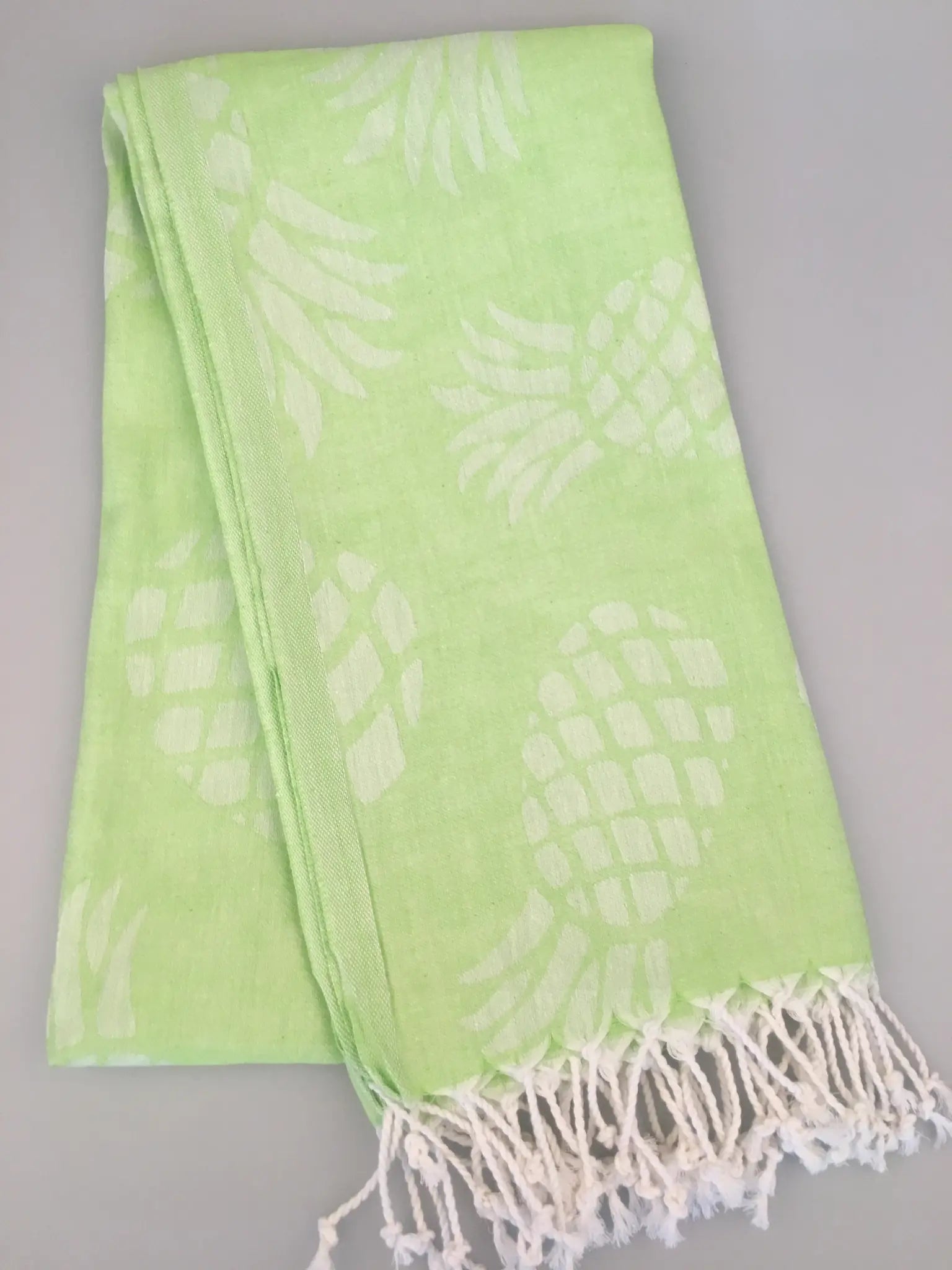 Light green Turkish towel with white pineapple jacquard pattern and fringed edges, folded view on a gray background.
