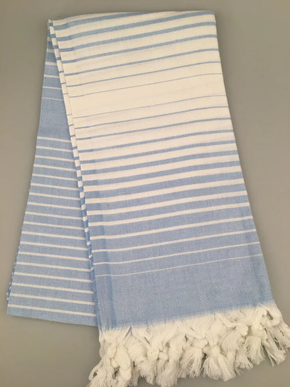 "Telmessos Turkish towel peshtemal in light blue and white stripes, fringed edges, folded on a gray background."