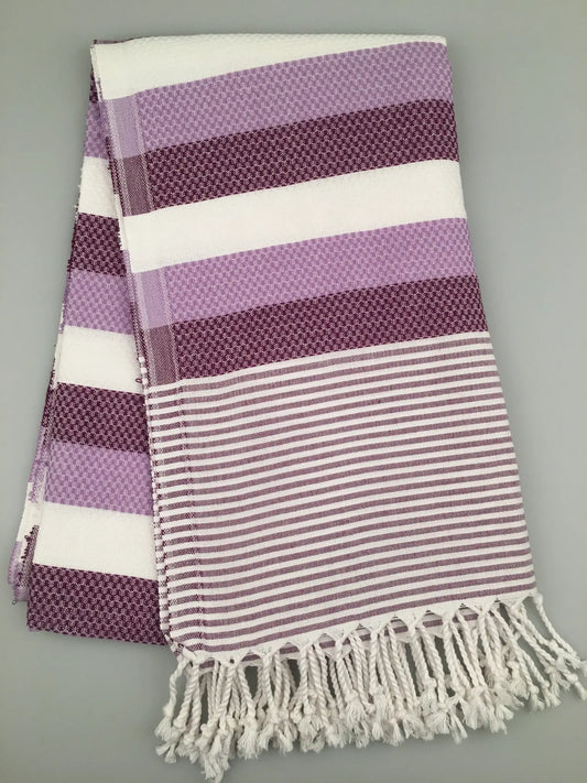 "Purple and white striped Turkish towel with textured patterns and fringed edges, laid flat on a gray background."