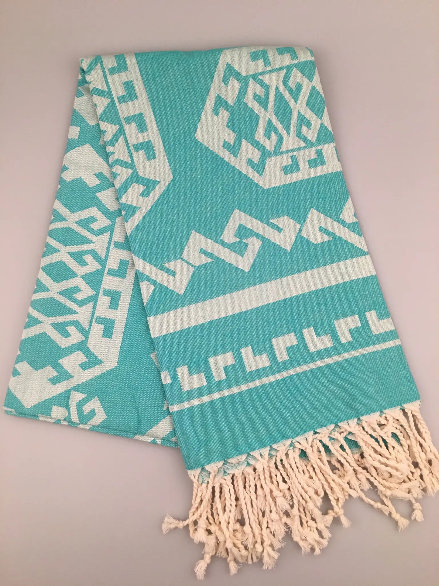 Turquoise Turkish towel with white jacquard pattern and fringed edges, laid flat on a light background.