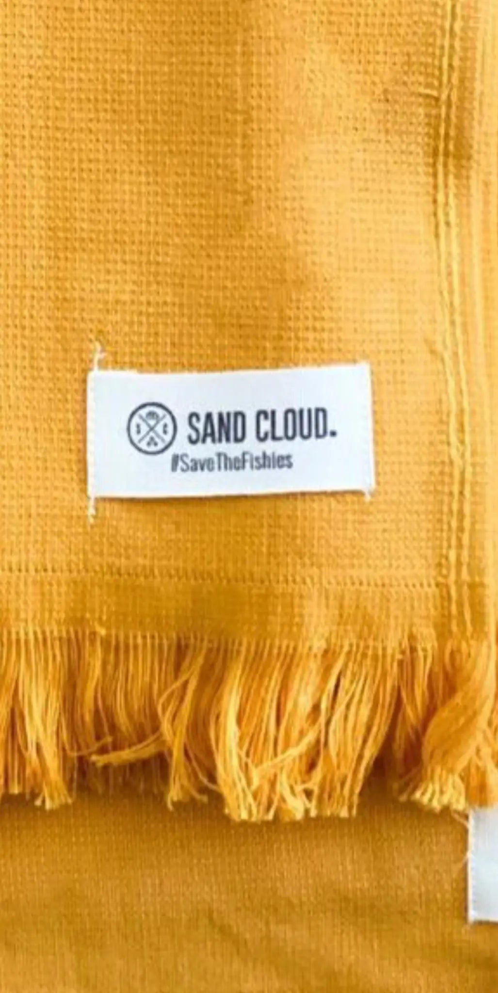 "Yellow cotton label with white text reading 'SAND CLOUD.' and hashtag '#SaveTheFishies,' featuring a fringed edge."