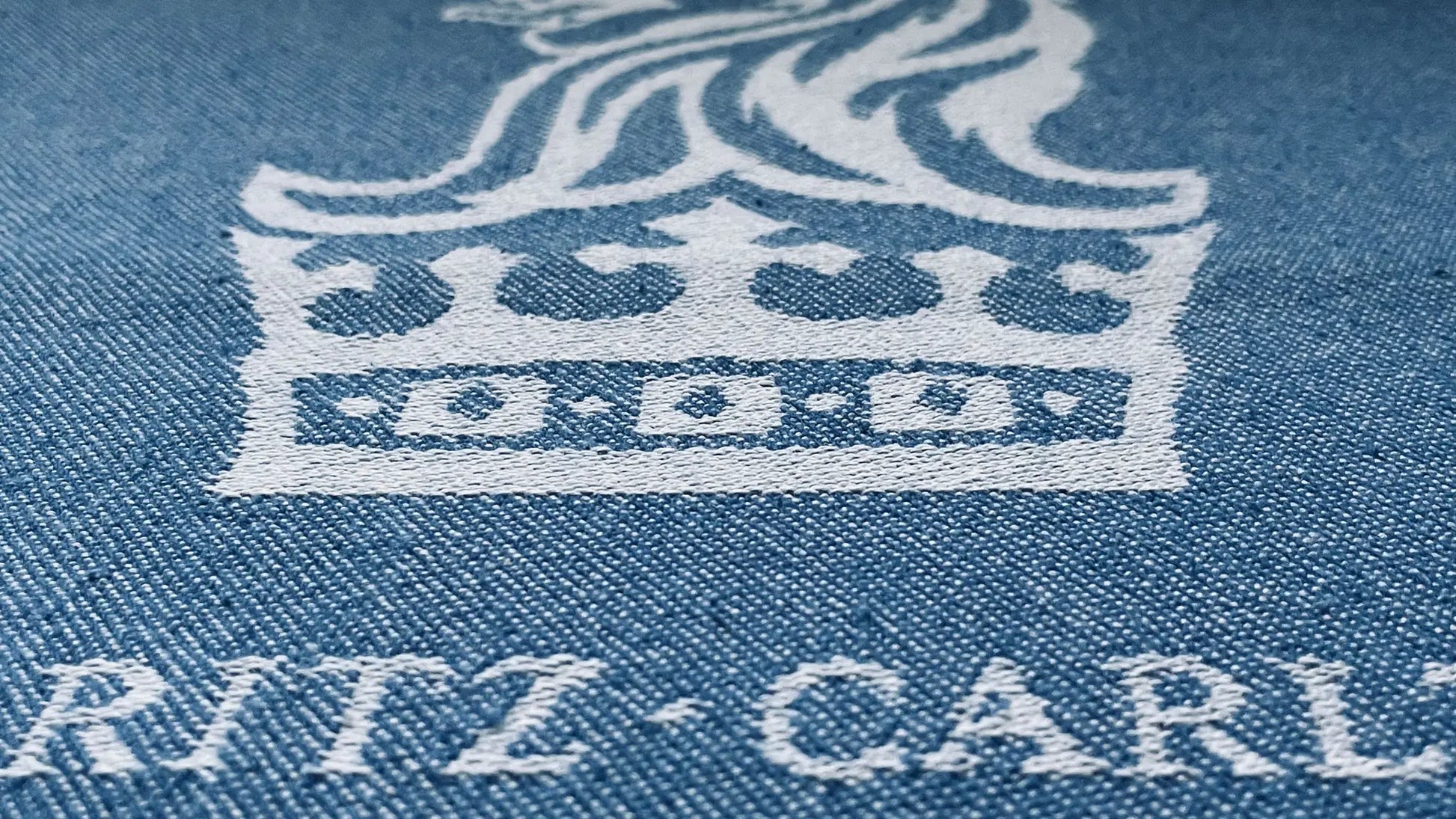 Light blue denim fabric with white embroidered crown logo and "RITZ-CARLTON" text beneath, close-up view.