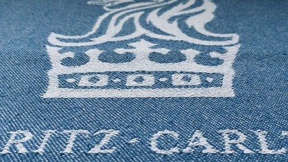 Light blue denim fabric with white embroidered crown logo and "RITZ-CARLTON" text beneath, close-up view.