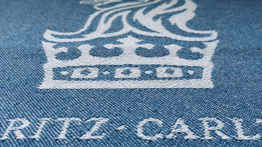 Light blue denim fabric with white embroidered crown logo and "RITZ-CARLTON" text beneath, close-up view.