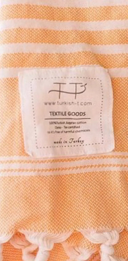 Orange and white striped cotton towel with a sewn label reading "TEXTILE GOODS" and a decorative fringe at the bottom.
