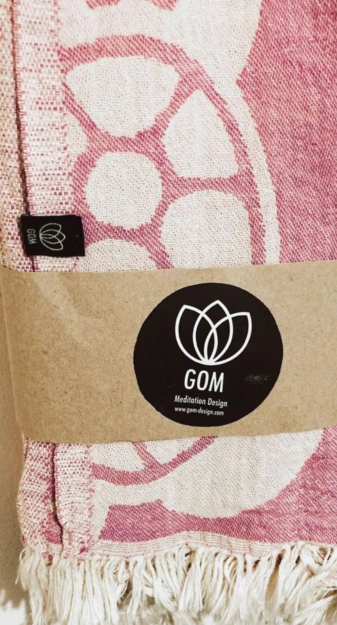 "Pink and cream cotton label with floral design, attached to a woven fabric with fringed edges, featuring a round logo."