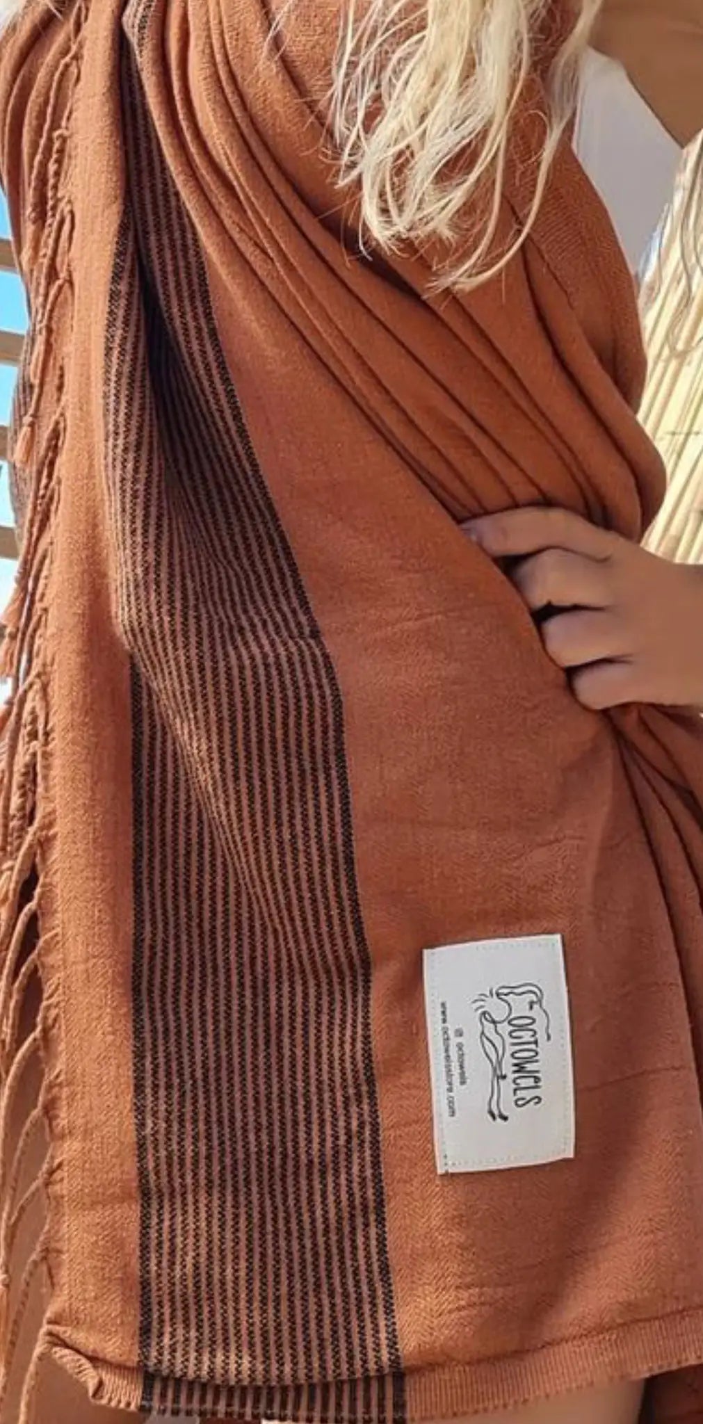 "Rust-colored cotton label with brown striped pattern and fringed edges, held by a person against a light background."