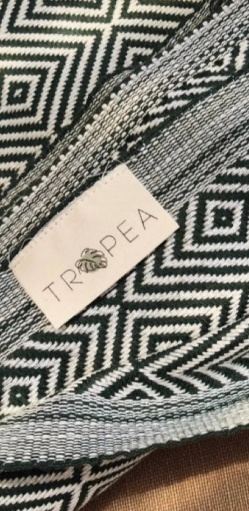 "White fabric label with black geometric pattern and 'TROPEA' text, stitched onto a patterned green textile."